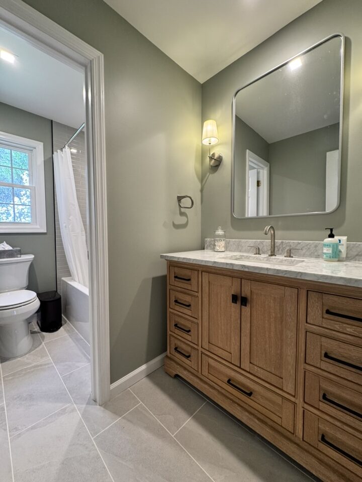 Bathroom vanity with sage green walls, natural oak cabinet, marble countertop, rounded mirror, and wall sconce
